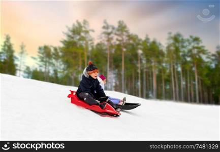 childhood, sledging and season concept - happy little kids sliding on sleds down snow hill over winter forest background. happy kids sliding on sleds down hill in winter