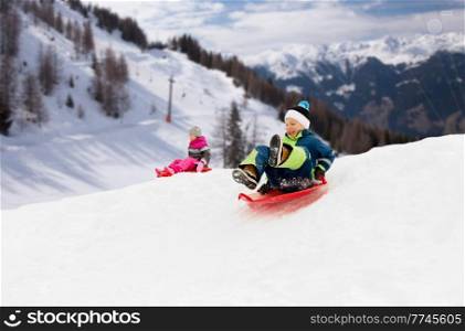childhood, sledging and season concept - happy little kids sliding on sleds down snow hill in winter over alps mountains on background. happy kids sliding on sleds down hill in winter
