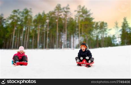 childhood, sledging and season concept - happy little kids sliding on sleds down snow hill in winter over snowy park or forest background. kids sliding on sleds down snow hill in winter
