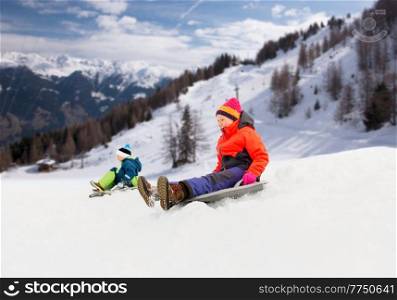 childhood, sledging and season concept - happy little kids sliding on sleds down snow hill in winter over alps mountains on background. happy kids sliding on sleds down hill in winter