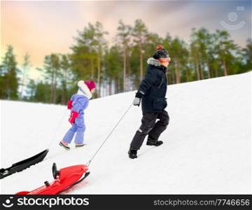 childhood, sledging and season concept - little children with sleds climbing snow hill in winter over snowy forest or park background. children with sleds climbing snow hill in winter