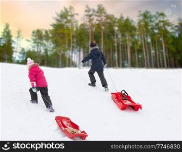 childhood, sledging and season concept - little children with sleds climbing snow hill in winter over snowy forest or park background. children with sleds climbing snow hill in winter