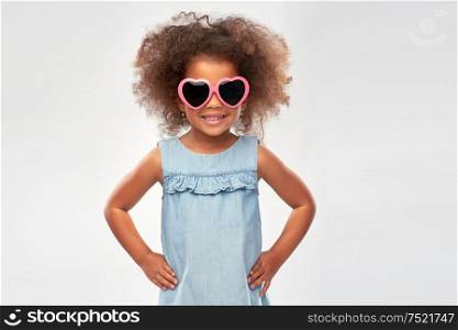 childhood, summer accessory and valentines day concept - happy little african american girl in heart shaped sunglasses over grey background. little african girl in heart shaped sunglasses