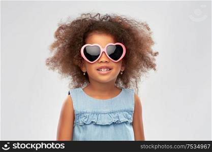 childhood, summer accessory and valentines day concept - happy little african american girl in heart shaped sunglasses over grey background. little african girl in heart shaped sunglasses
