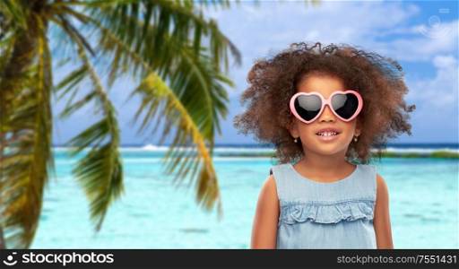 childhood, summer accessory and valentines day concept - happy little african american girl in heart shaped sunglasses over tropical beach background in french polynesia. little african girl in sunglasses on beach