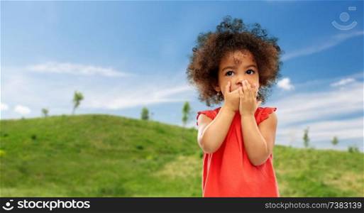 childhood, summer and emotions concept - confused little african american girl covering mouth by hands over green meadow and blue sky background. confused african american girl covering mouth