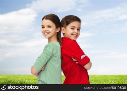 childhood, summer and people concept - happy smiling boy and girl standing back to back over blue sky and grass background