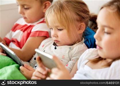 childhood, technology and family concept - little kids with tablet pc computer and smartphone in bed at home. kids with tablet pc and smartphone in bed at home