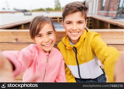 childhood, technology and people concept - happy children or brother and sister sitting on wooden street bench outdoors taking selfie. children sitting on street bench and taking selfie
