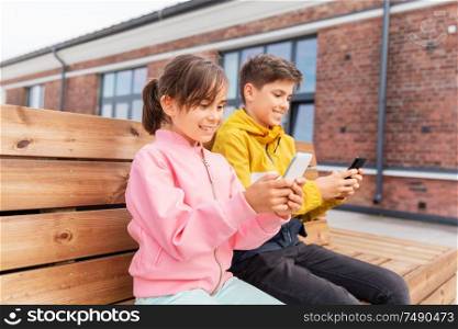 childhood, technology and people concept - happy children or brother and sister with smartphones sitting on wooden street bench outdoors. children with smartphones sitting on street bench