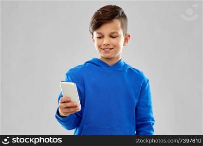 childhood, technology and people concept - smiling boy in blue hoodie using smartphone over grey background. smiling boy in blue hoodie using smartphone