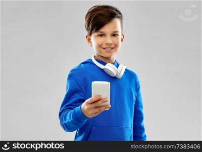 childhood, technology and people concept - smiling boy in blue hoodie with headphones on neck using smartphone over grey background. smiling boy in blue hoodie using smartphone
