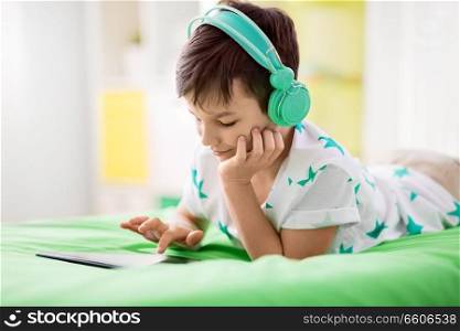 childhood, technology and people concept - smiling boy with tablet pc computer and headphones lying on bed at home. smiling boy with tablet pc and headphones at home