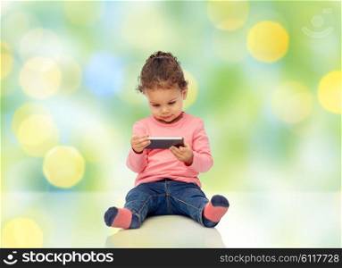 childhood, technology and people concept - smiling little african american baby girl playing with smartphone and sitting on floor over green summer light background