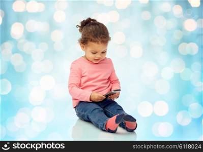 childhood, technology and people concept - smiling little african american baby girl playing with smartphone and sitting on floor over blue holidays lights background