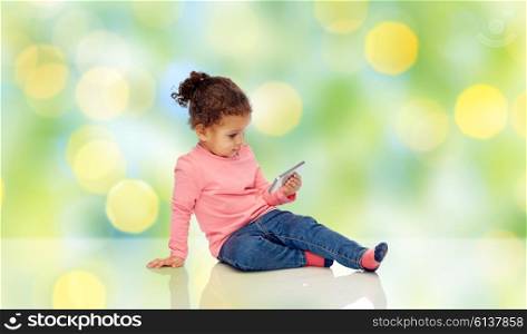 childhood, technology and people concept - smiling little african american baby girl playing with smartphone and sitting on floor over green summer holidays lights background