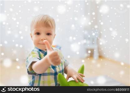childhood, toys and people concept - happy little baby boy playing with ride-on toy horse and showing thumbs up at home over snow