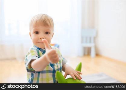childhood, toys and people concept - happy little baby boy playing with ride-on toy horse and showing thumbs up at home