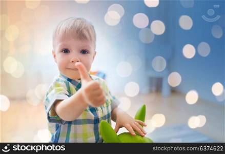 childhood, toys and people concept - happy little baby boy playing with ride-on toy horse and showing thumbs up at home. happy baby boy playing with toy showing thumbs up