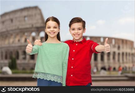 childhood, travel, tourism, gesture and people concept - happy smiling boy and girl hugging and showing thumbs up over coliseum in rome