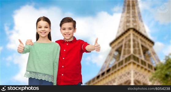childhood, travel, tourism, gesture and people concept - happy smiling boy and girl hugging and showing thumbs up over paris eiffel tower background