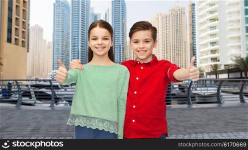 childhood, travel, tourism, gesture and people concept - happy smiling boy and girl hugging and showing thumbs up over dubai city street background