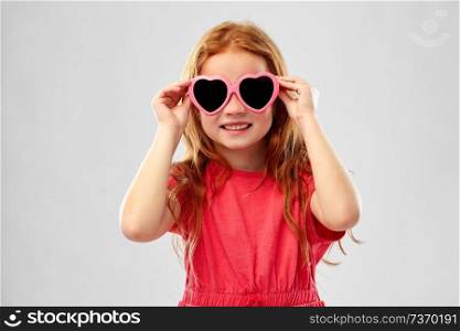 childhood, valentine&rsquo;s day and summer concept - smiling red haired preteen girl in heart shaped sunglasses over grey background. smiling red haired girl in heart shaped sunglasses