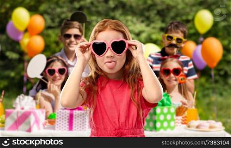 childhood, valentines day and summer concept - naughty red haired girl with heart shaped sunglasses showing tongue over birthday party at park. naughty red haired girl in heart shaped sunglasses