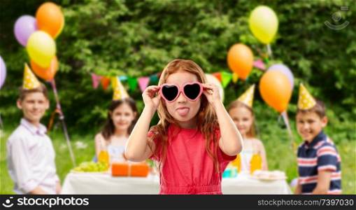 childhood, valentines day and summer concept - naughty red haired girl with heart shaped sunglasses showing tongue over birthday party at park. naughty red haired girl in heart shaped sunglasses