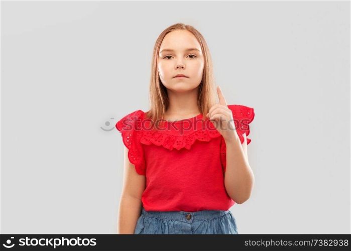 childhood, warning and attention concept - beautiful serious girl in red shirt pointing finger up over grey background. serious girl in red shirt pointing finger up
