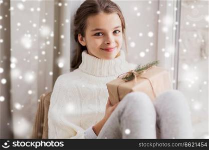childhood, winter holidays and people concept - happy beautiful girl with christmas gift sitting at home over snow. girl with christmas gift sitting at home