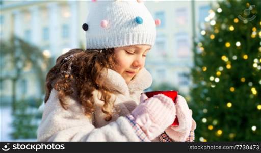 childhood, winter holidays and season concept - happy little girl with cup of hot tea outdoors over christmas tree lights on background. little girl with cup of hot tea on christmas