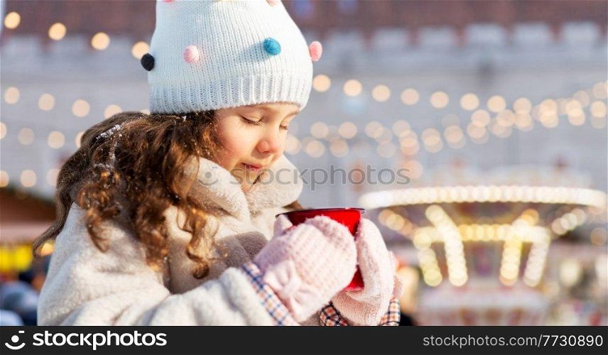 childhood, winter holidays and season concept - happy little girl with cup of hot tea over christmas market or amusement park background. little girl with cup of tea over christmas market