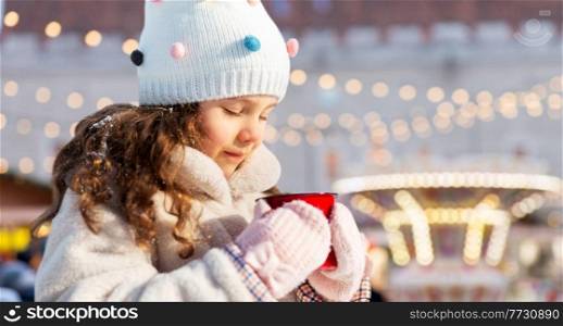 childhood, winter holidays and season concept - happy little girl with cup of hot tea over christmas market or amusement park background. little girl with cup of tea over christmas market