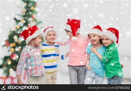 childhood, winter holidays, friendship and people concept - group of happy smiling little children in santa hats hugging over christmas tree and snow
