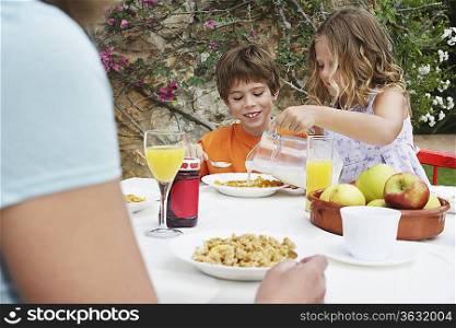 Children (5-6) at table, girl pouring milk onto cereal