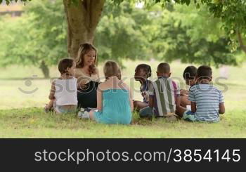 Children and education, young woman at work as educator reading book to boys and girls in park