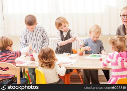 Children and tutor are painting with a brush and watercolors on paper in the kindergarten. Children are painting