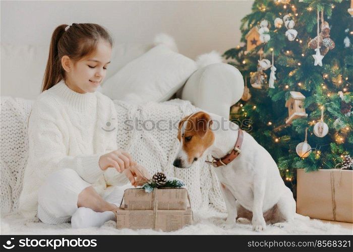 Children, animals and winter holidays concept. Charming little European girl unpacks gift box, poses on floor together with pedigree puppy, prepare for New Year and Christmas celebration. Festive time