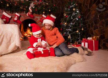 Children are sitting near fireplace and christmas tree with gift boxes.