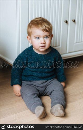 Children concept. Little small infant with appealing blue eyes, plump cheeks and blonde hair sits on floor, teacher to go by himself, wears sweater and trousers. Small child with serious expression