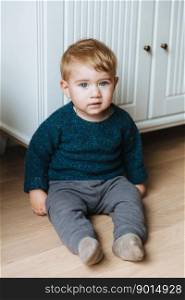 Children concept. Little small infant with appealing blue eyes, plump cheeks and blonde hair sits on floor, teacher to go by himself, wears sweater and trousers. Small child with serious expression