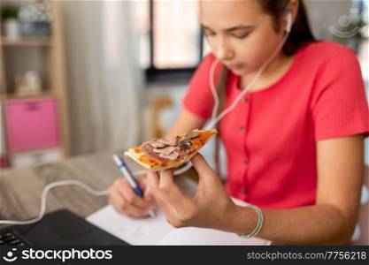 children, education and distant learning concept - close up of teenage student girl in earphones with laptop computer eating pizza and writing to notebook at home. student girl in earphones eating pizza at home