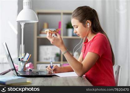 children, education and distant learning concept - teenage student girl in earphones with laptop computer and pizza writing to notebook at home. student girl in earphones with pizza at home