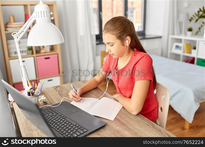 children, education and distant learning concept - teenage student girl in earphones with laptop computer writing to notebook at home. student girl in earphones learning at home