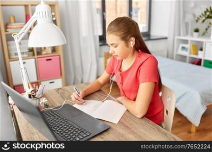 children, education and distant learning concept - teenage student girl in earphones with laptop computer writing to notebook at home. student girl in earphones learning at home