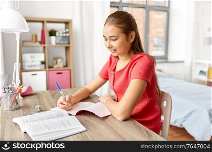 children, education and learning concept - happy smiling student teenage girl with book writing to notebook at home. student girl with book writing to notebook at home