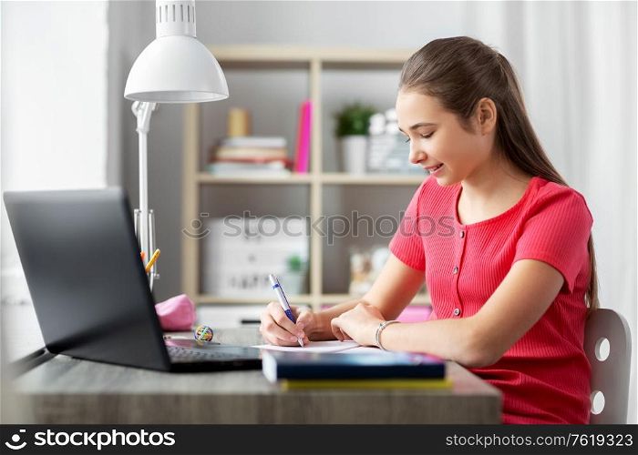 children, education and learning concept - happy smiling teenage student girl with laptop computer writing to notebook at home. teenage student girl writing to notebook at home