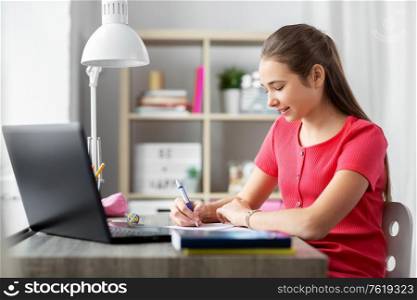 children, education and learning concept - happy smiling teenage student girl with laptop computer writing to notebook at home. teenage student girl writing to notebook at home