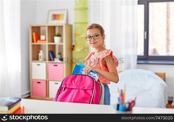 children, education and learning concept - happy student girl in glasses packing school bag at home. student girl in glasses packing school bag at home